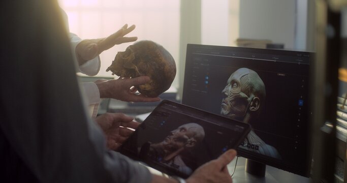 Modern archaeological lab: Two scientists analyze fossil skull, discuss three dimensional model and visualization of prehistoric extinct human head displayed on digital tablet and computer screen.