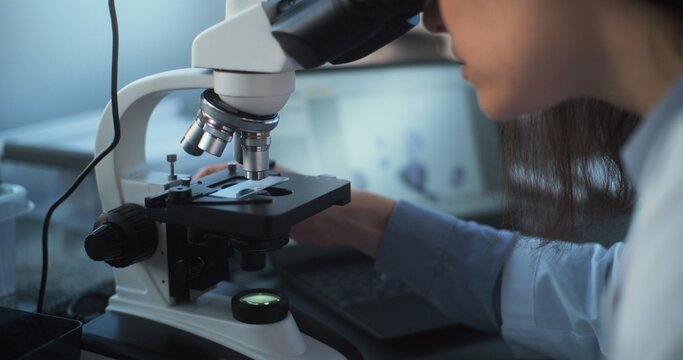 Close up of young scientist using microscope, analyzing sample in archaeological or medical science laboratory, looking at laptop screen. Professional microbiologist works with advanced equipment.