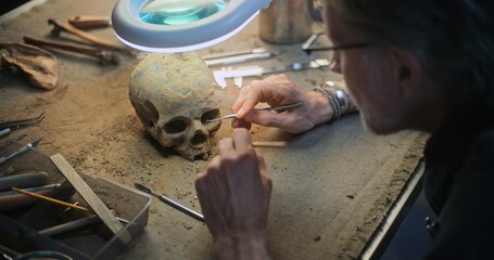 Mature archaeologist cleans fossil skull under lamp using professional tools and brush. Scientist studies remains of extinct prehistoric human, works in archaeological laboratory. Close Up. Zoom In.