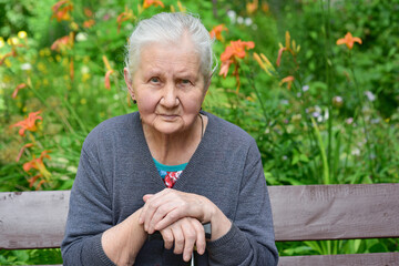 Portrait of a sad elderly woman, 80 years old, with a cane in her hands, sitting on a bench in the park.