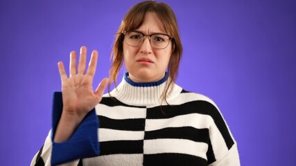 Young sad unhappy woman with brown hair wearing glasses and sweater shaking head NO holding up had in STOP gesture, isolated on purple background with copy space.