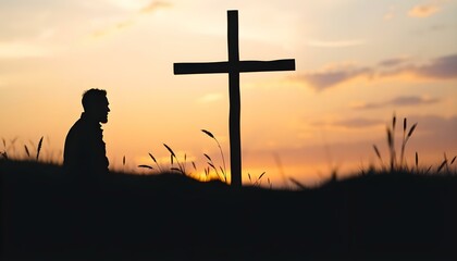 Silhouette of man kneeling near the wooden Christian cross on a nature meadow grass field outdoor at sunset
