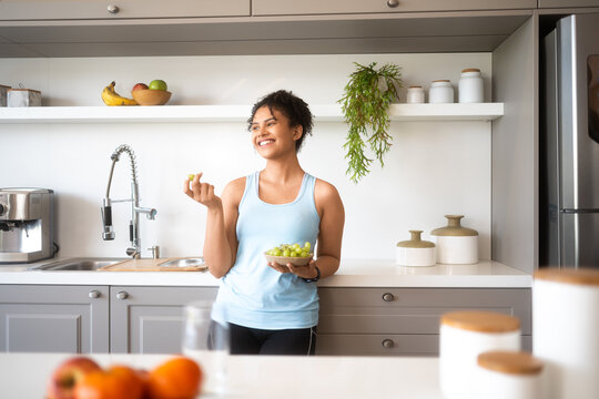 Happy latin woman eating green grapes and leaning in kitchen counter at home.. - Powered by Adobe