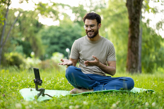 Young man conducts online yoga session in park, demonstrating mindfulness and relaxation techniques via smartphone