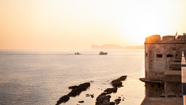 Partial view of the ancient defensive wall of the coastal city of Alghero in front of an intense sunset over the sea with two boats returning to the port on the island of Sardinia, Italy