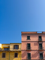 The facades of two adjacent buildings, one red and one yellow, create a perfect color triad against the blue sky. Located in Bosa, Italy, with Italian-style windows and Venetian shutters
