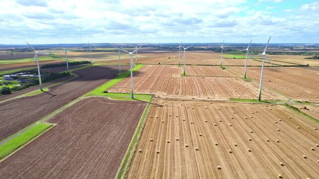 With a bird's-eye view, we observe the wind turbines elegantly turning in a Lincolnshire farmer's freshly harvested field, complemented by the presence of golden hay bales.