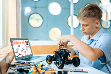 Blonde hair boy carefully using pliers fixing robotics car while learning technology in STEM class. On table put laptop, controller, electric wire, battery charger, and robotic vehicle. Edification.