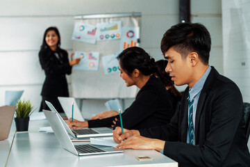 Asian business people listening to presentation from businesswoman who stands in front of group meeting room at international business conference. Multicultural corporate strategy concept. uds