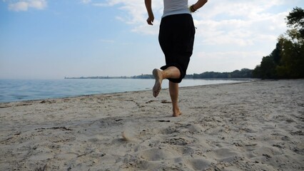 Bare feet of adult woman jogging on sand at the beach with seascape at background. Female legs running along sea shore. Happy lady spending time near ocean coast. Concept of summer vacation