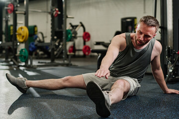 Tired and sweaty athlete sitting on floor and doing stretching after intense workout in gym