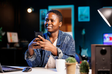 While sitting at her home desk, african american self employed individual checks cell phone for messages. A young black woman using mobile device to chat on social media.