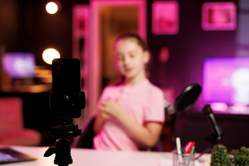 Focus on phone attached to tripod filming girl in home studio talking about day at school with children watching her. Kid in blurry background recording content for online channel with smartphone