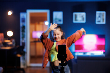 Focus on phone attached to tripod filming child taking part in viral dance trend after seeing favorite celebrities doing it. Talented youngster in blurry background does trendy dancing challenge
