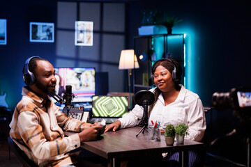 Cheerful media stars recording a podcast in home studio with professional filming equipment for broadcast on live streaming platform. Blogger interviewing celebrity, filming the encounter with camera.