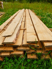 A pile of wood is stacked in a field. The wood is brown and has a rough texture. The grass is green and the field is open. Construction material for building. Natural product
