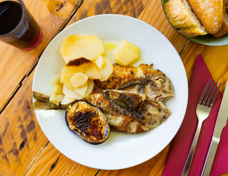 There is plate with lunch on simple wooden table. Dish is made from fried fish carcass of lean dorado, baked potato vegetables and several round pieces of eggplant