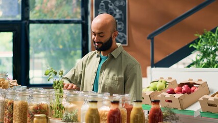 Young man explores eco friendly store, browsing through variety of fresh organic pantry products with reusable packaging. Middle eastern client looking at pasta and grains in jars.