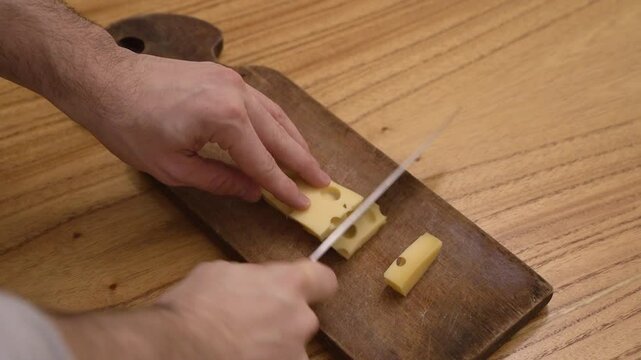 Top view of a caucasian man hands slicing a gruyere cheese with a kitchen knife and a wooden cutting board