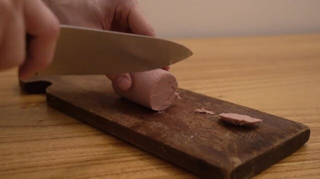 Closeup view of a man slicing a german leberwurst with a sharp kitchen knife and a wooden cutting board