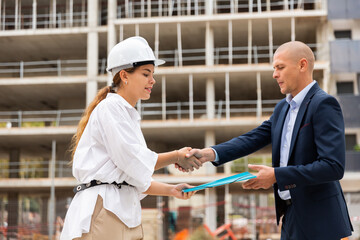 Young woman real estate manager giving paper-folder with documentation to man who have purchased apartment in new multistory house.