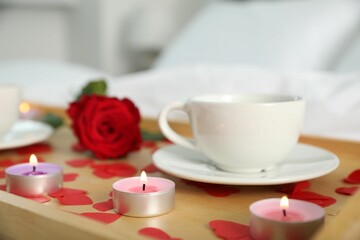 Wooden tray with burning candles, rose, cup of drink and red paper hearts on bed indoors, closeup