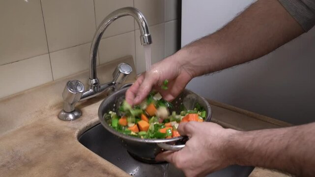 Preparing a homemade soup. Closeup view of a caucasian man washing sliced vegetales in a colander under the tap water in the kitchen sink
