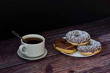 A cup of hot coffee on a saucer with a spoon and a few donuts in chocolate icing on a wooden table.
