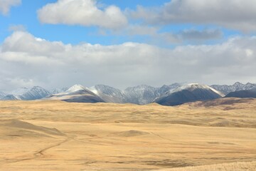 Field roads bending around high hills cross the yellowed steppe at the foot of snow-capped mountains on a sunny autumn day.