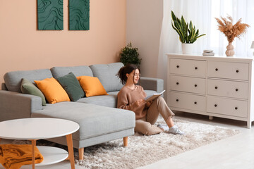 Young woman in eyeglasses reading book on floor near comfortable sofa in living room