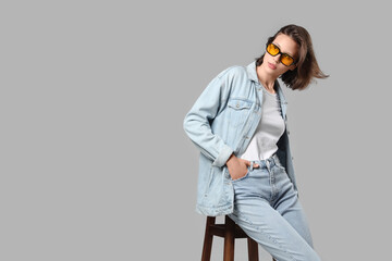 Beautiful young woman in stylish denim clothes sitting on chair against grey background