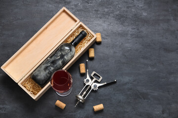 Box with bottle of wine, glass, corks and opener on black grunge background