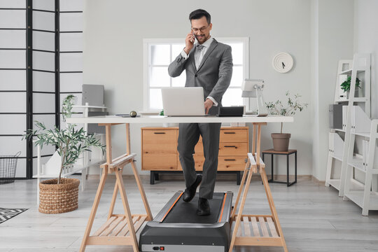 Handsome businessman talking by mobile phone on treadmill at table in office - Powered by Adobe