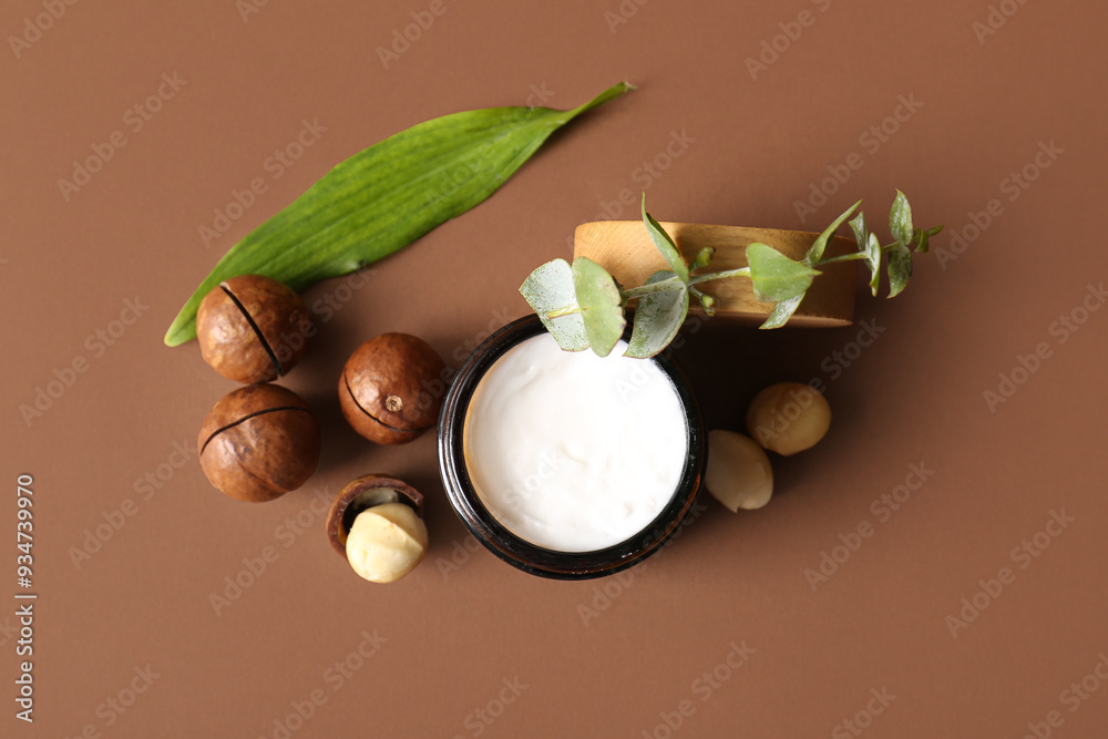 Wall mural Macadamia nuts, green leaves and jar of cosmetic product on brown background, closeup