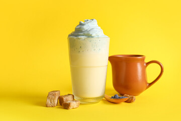 Glass of blue dalgona matcha latte, jug and brown cane sugar cubes on yellow background