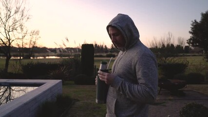 Argentinian traditional infusion drink. View of a man pouring hot water into a mate containing yerba mate herbs and drinking through the metal straw, in the garden at sunset 