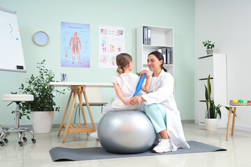 Female osteopath and little patient with elastic band doing exercises on fit ball in clinic