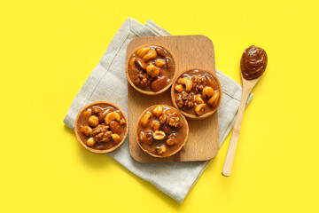 Tasty tarts with nuts and caramel on wooden board against yellow background