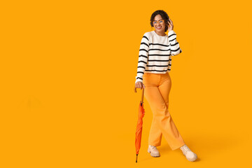 Beautiful young happy African-American woman in autumn outfit and headphones with umbrella on orange background