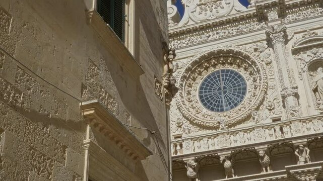 Intricate facade of a historic baroque church in lecce, puglia, italy, featuring detailed stone carvings and a large circular stained-glass window under a clear blue sky.