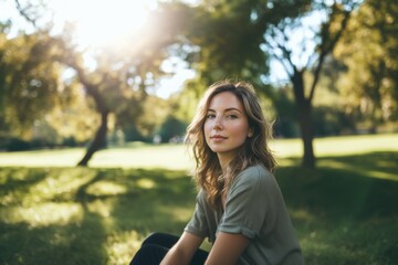 A candid portrait of a White woman in her early 30s, with natural makeup and a casual outfit, sitting in a sunny, open park 