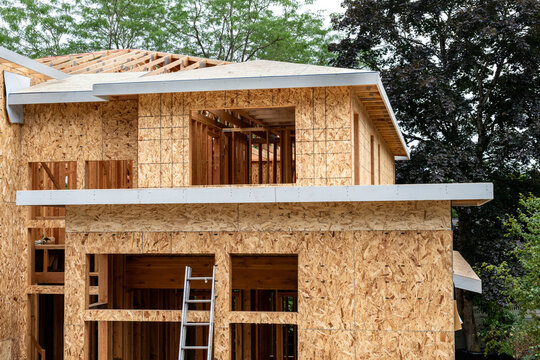 New large residential home construction in framing stage, with exterior walls covered in OSB sheathing, roof sheathing installation started over wood rafters, cloudy summer day
