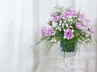 Purple flowers and green leaves in a clear glass vase, white curtain background. Leave the copy space.