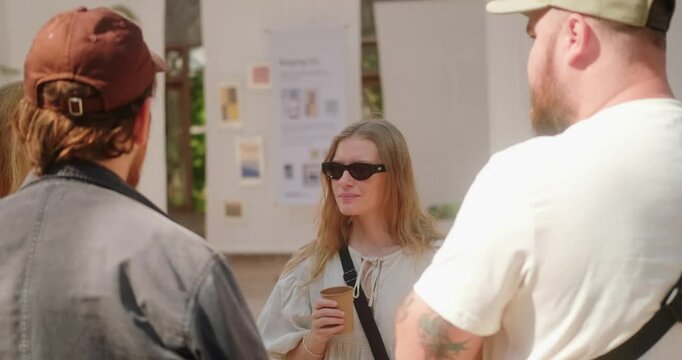 A group of four friends, men and women, stand at an outdoor contemporary art exhibition and discuss their impressions. A woman holds a paper cup of coffee and smiles. Joint leisure time of art lovers.