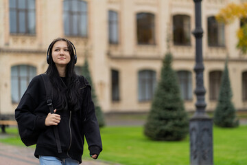 A girl student goes to campus with a backpack on her shoulders and listens to music in headphones