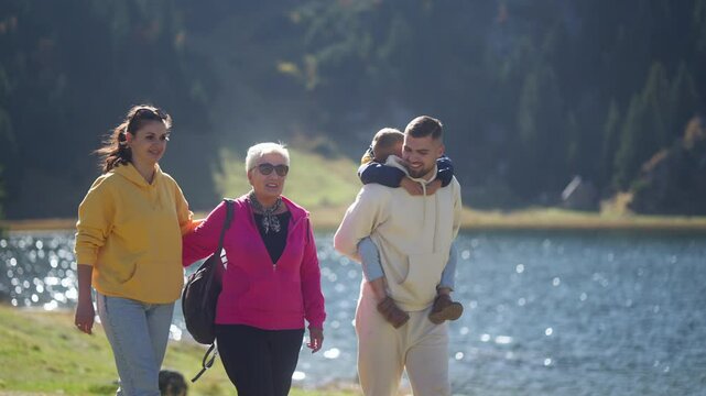 Joyful Father Carrying Son on His Back During Lakeside Walk with Wife and Mother-in-law on Sunny Day. Concept of Fatherhood, Fun, Connection, Bonding, Love, Happy Moments, Togetherness, Relationship.