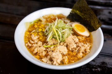 A bowl of Miso Japanese ramen noodles, featuring half a boiled egg, corn, seaweed, and shredded meat in a savory broth, served in a white bowl on the black wood floor.