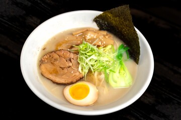 This image showcases a Tonkotsu ramen bowl of traditional Japanese ramen with slices of pork, halved boiled egg, seaweed, green onions, and other toppings, served in a white bowl.