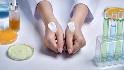 The advertising photo was taken from top view, two scientist hands placed on white lab table with white cream streaks of cosmetic applied on back of hand, surrounded by lab glassware contain essence