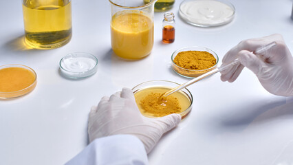 Close up high angle view at the white lab table top, a scientist is using a glass rod to stir turmeric powder and yellow essence in petri dish, next to glassware contain experiment composition